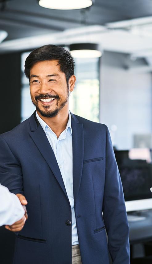 Three professionals in business attire stand in an office; two are shaking hands while one holds a tablet and observes, all appearing engaged in conversation.