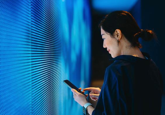 A woman gazes at her phone while standing against a plain wall, appearing engaged in her device.